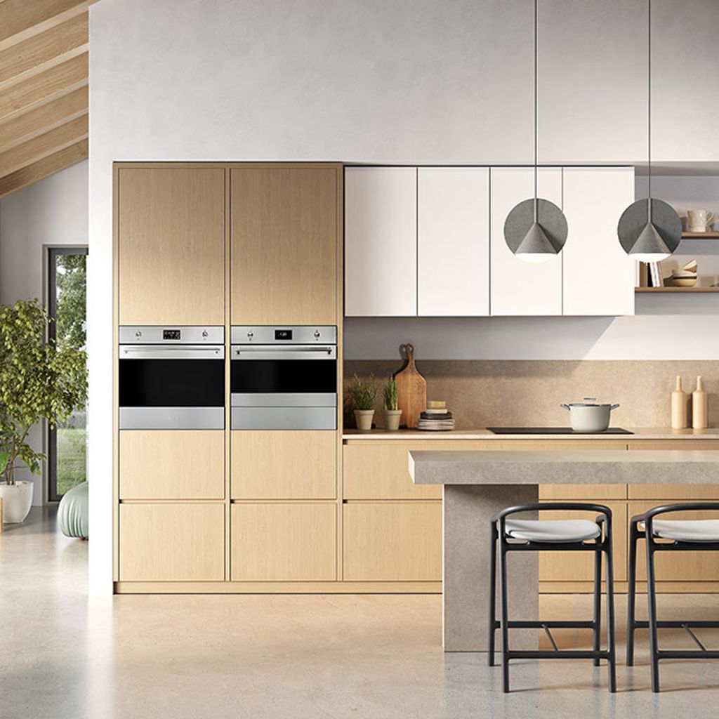 A bright and modern kitched with light wooden units, and two integrated Smeg ovens (stainless steel) positioned side-by-side across two floor-to-ceiling columns of kitchen units