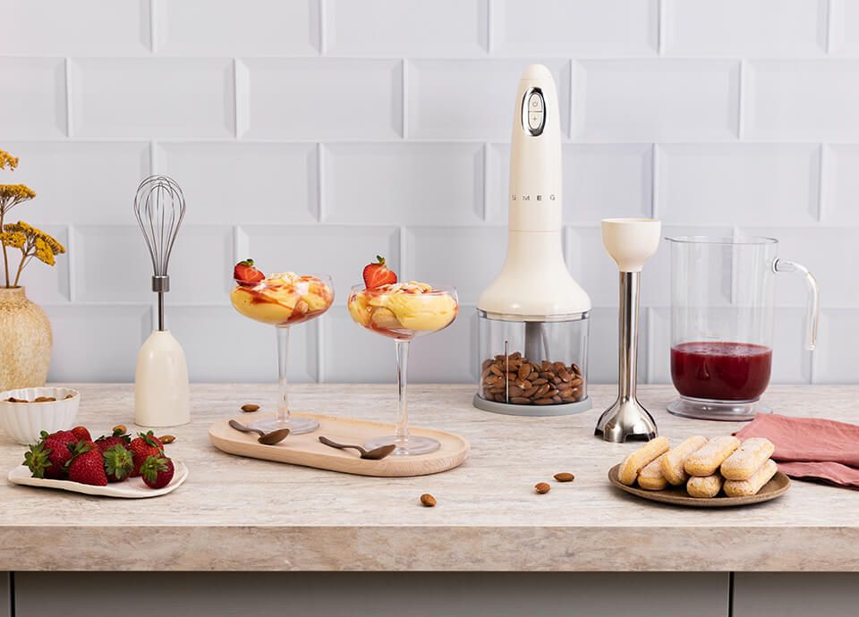 Frullatore ad immersione rosso con accessori e, sul piano lavoro, frutta e verdura feschi