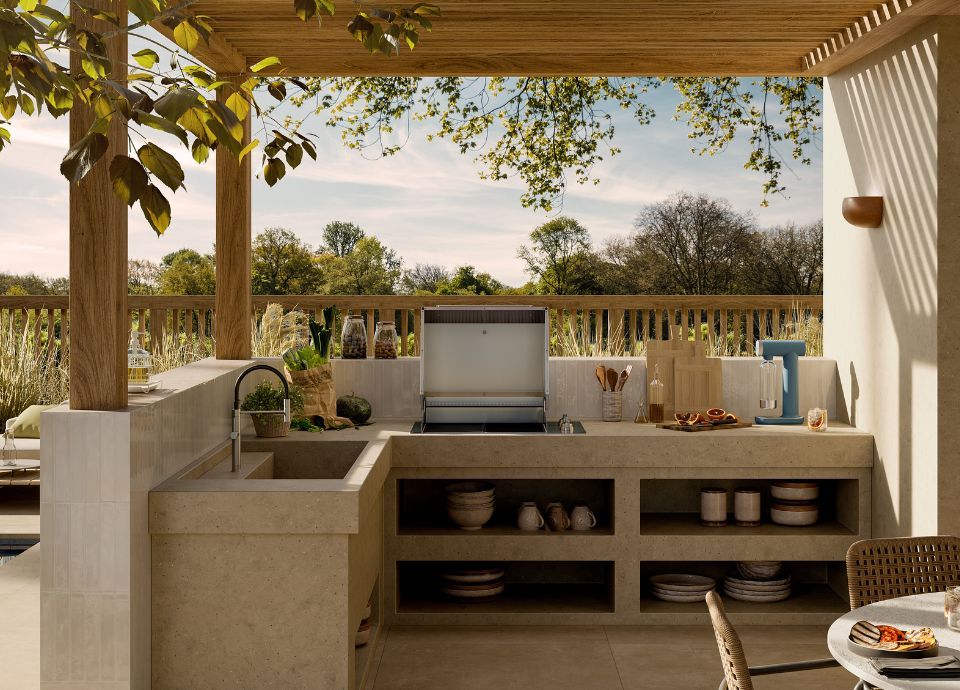 Outdoor kitchen under a wooden pergola. On the light stone countertop: built-in stainless steel barbecue with open lid, sink with chrome faucet, utensils and jars. Lower shelves with plates and bowls. Background shows a green garden with trees. Foreground includes a partially set table.