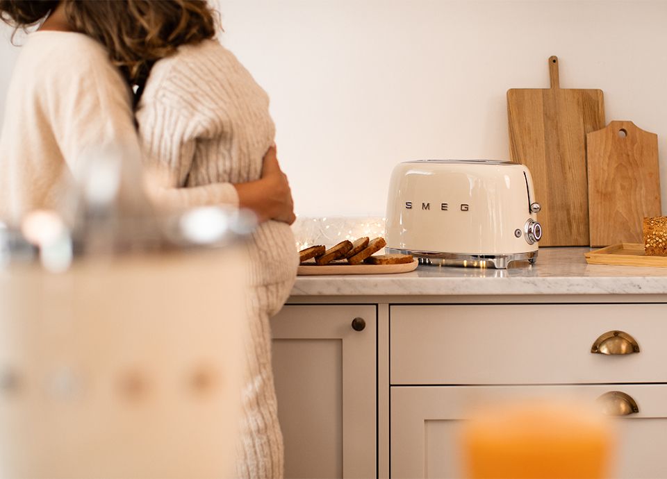 Mother and daughter hug in the kitchen, wearing light-colored sweaters. In the foreground, there is a SMEG citrus juicer, and on the countertop, a cream-colored SMEG toaster, a plate with slices of toasted bread, and wooden cutting boards in the background.