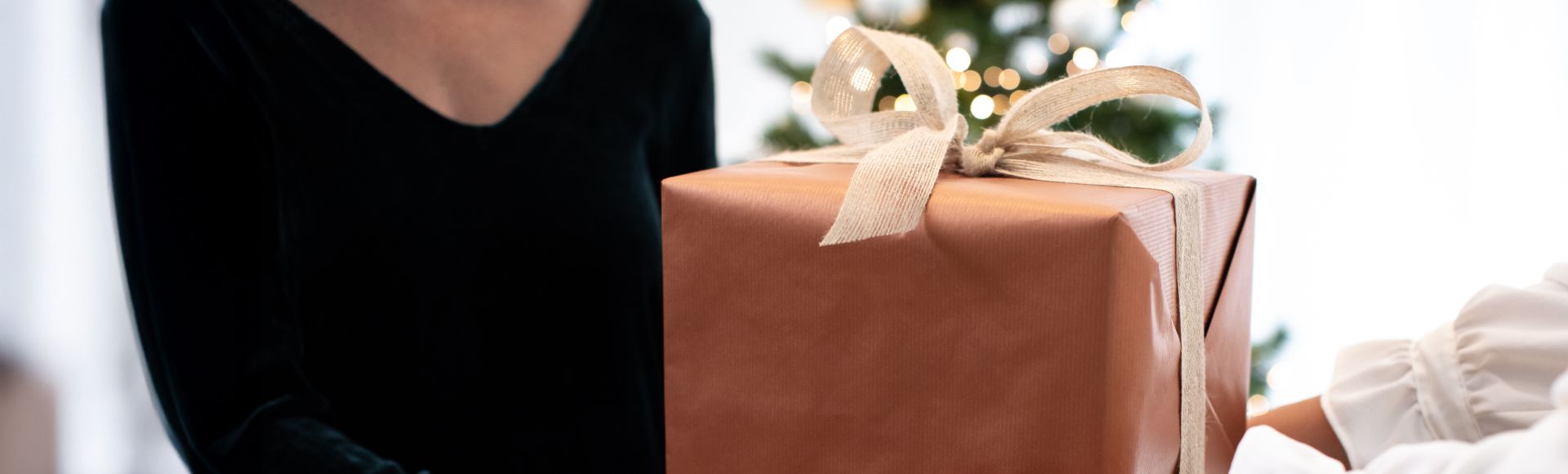 Two people exchange a large rectangular gift wrapped in brown paper and tied with a beige ribbon. A Christmas tree with warm lights is visible in the background.