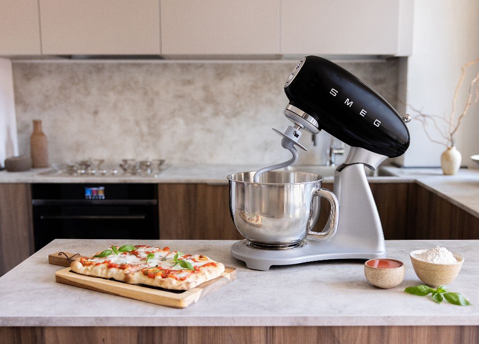 Black SMEG stand mixer with a metal body and stainless steel bowl placed on the kitchen countertop. Next to it, on the counter, there is a pizza tray topped with tomato, mozzarella, and fresh basil leaves. There are also three small bowls: one with white flour, one with tomato, and one with basil leaves. In the background, you can see a light wood kitchen with beige wall cabinets, a built-in SMEG oven, a stainless steel SMEG hob and some decorative items.