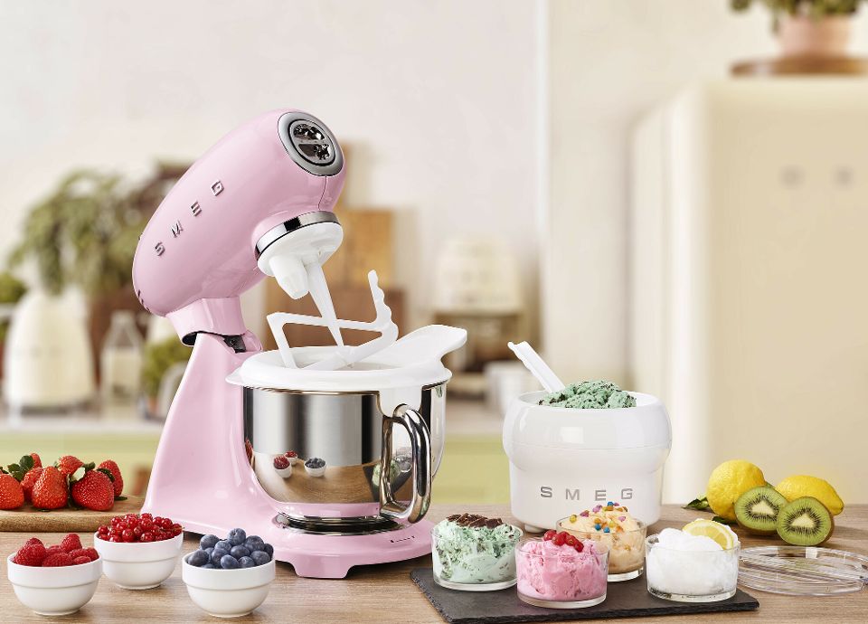 On a light-colored kitchen countertop, there is a pink SMEG Stand Mixer with ice cream accessories and a SMEG ice cream container next to it. Fresh fruit, such as strawberries, kiwis, lemons, and small jars of berries, can also be seen on the kitchen countertop. There are also four small glass bowls filled with freshly made ice cream, each with a different flavor. In the background there are other SMEG kitchen appliances, placed on a light-colored countertop of a bright kitchen. The room is tidy and bright.