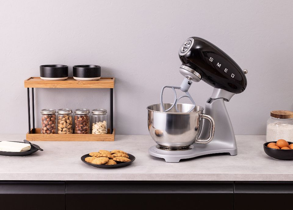 On a light-colored countertop in a black kitchen, there is a black SMEG Stand Mixer with stainless steel body, several glass jars filled with dried fruit resting on a small wooden shelf, a plate of cookies, a plate of butter, a jar of flour, and some eggs in a dark bowl. The room is tidy and modern.