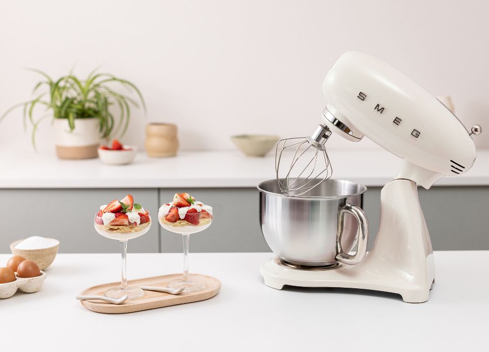 On a light-colored kitchen countertop, there is a white SMEG Stand Mixer. Next to the SMEG Stand Mixer, there are two desserts with strawberries on a wooden tray, some eggs and sugar in a container. In the background, there is a light-colored kitchen countertop with some little plants and strawberries in a pot. The room is tidy and bright.