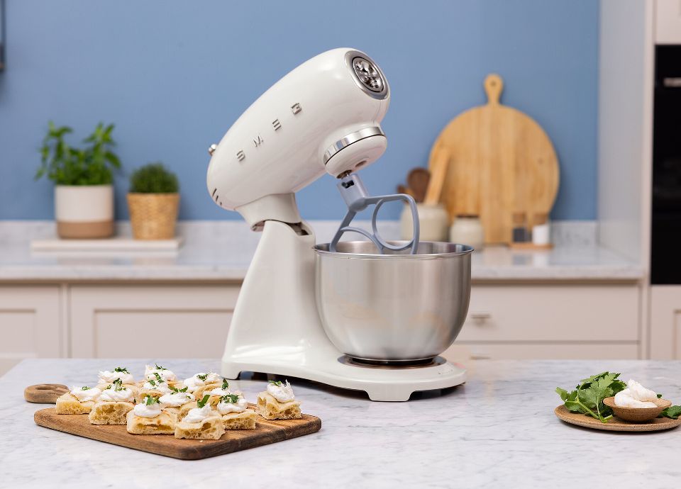 On a marble kitchen countertop there is a white SMEG Stand Mixer. Next to the SMEG Stand Mixer there are some appetizers and a small bowl with some cheese and parsley. In the blue background, there is a light-colored kitchen countertop with some plants in a pot, wooden cutting boards leaning against the wall, salt and pepper in a jar, and ladles. The room is tidy and bright.