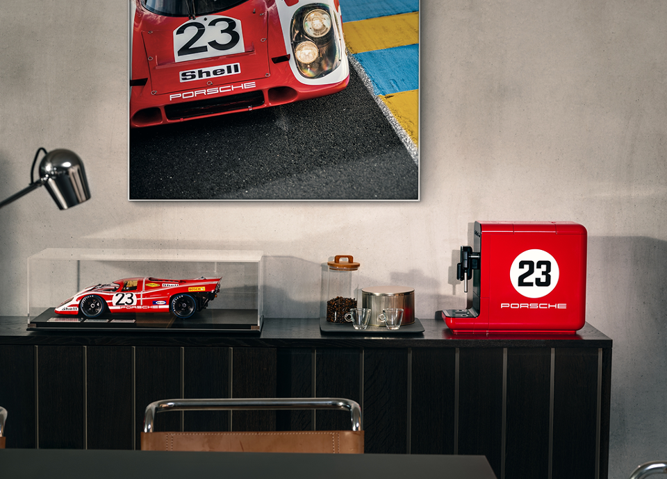 BCC12 917 Salzburg Porsche x SMEG Espresso Coffee Machine with the icon number 23 on the side. The machine is placed on a cabinet in an office setting. Next to it, there is a scale model and a framed picture of the 917 Salzburg race car, famous for winning Le Mans in 1970.