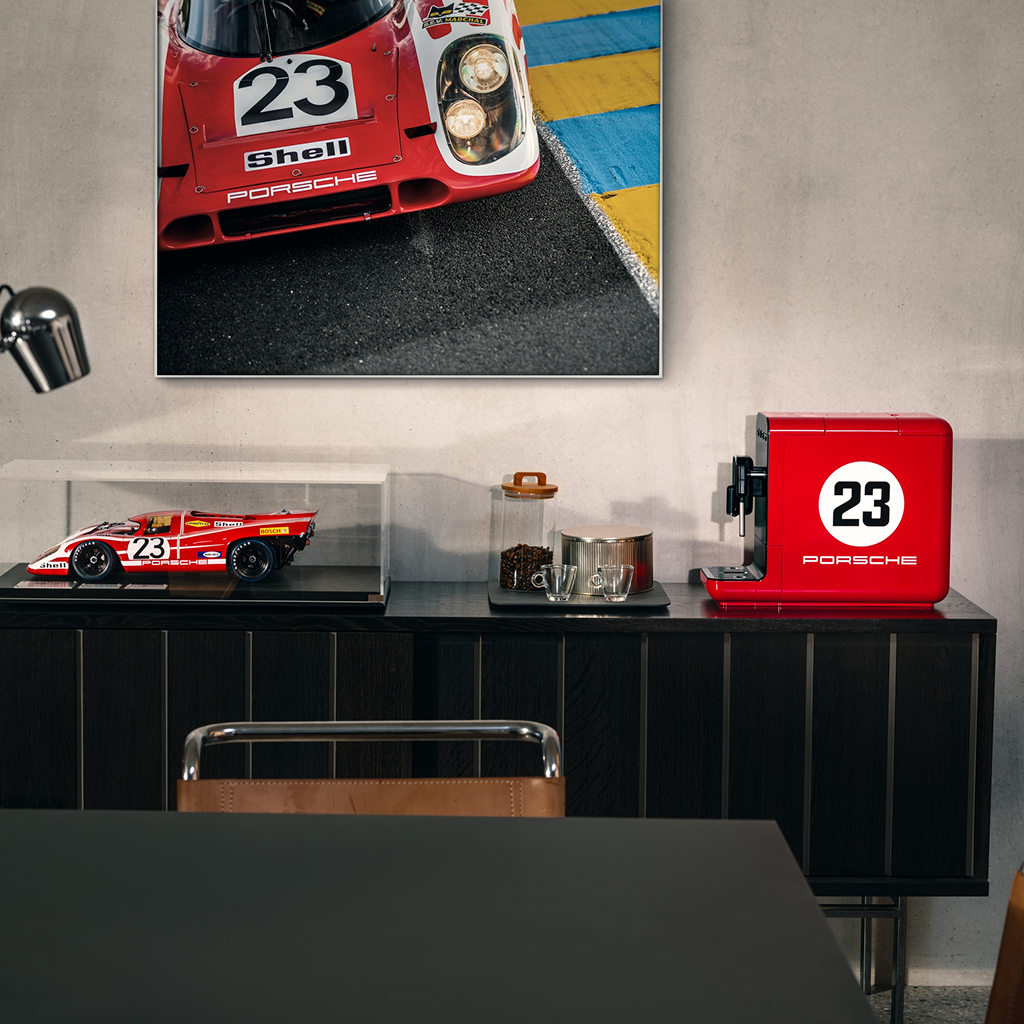 BCC12 917 Salzburg Porsche x SMEG Espresso Coffee Machine with the icon number 23 on the side. The machine is placed on a cabinet in an office setting. Next to it, there is a scale model and a framed picture of the 917 Salzburg race car, famous for winning Le Mans in 1970.