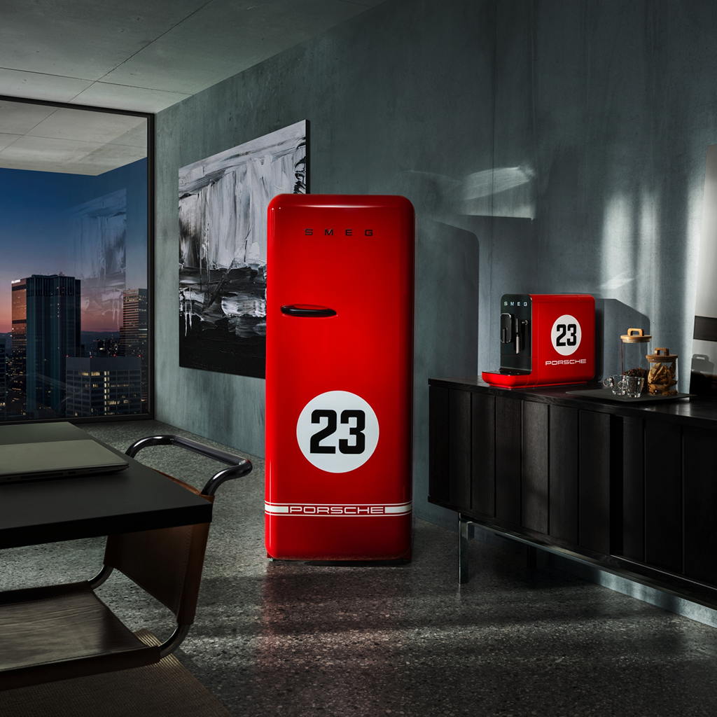 A modern, stylish interior with a large window revealing a cityscape at dusk. The room features sleek dark furniture and minimalist decor. Prominently displayed are a red SMEG refrigerator and a red coffee machine, each marked with a white circle containing the number "23" and "PORSCHE" logo. The refrigerator has the brand name "SMEG" in bold letters at the top. Behind it, a black and white abstract painting adds contrast to the scene. The design emphasizes bold colors, clean lines, and strong visual contrast, ensuring legibility and accessibility for users with visual impairments.
