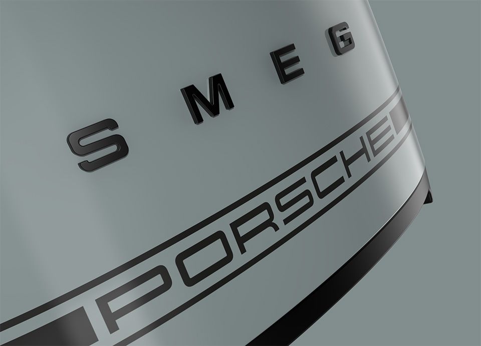 Detail of the front part of the toaster in colour shade green. The front of the toaster features the brand name "SMEG" in bold uppercase letters, and below it, a second logo reads "PORSCHE", indicating a collaboration between the two brands.